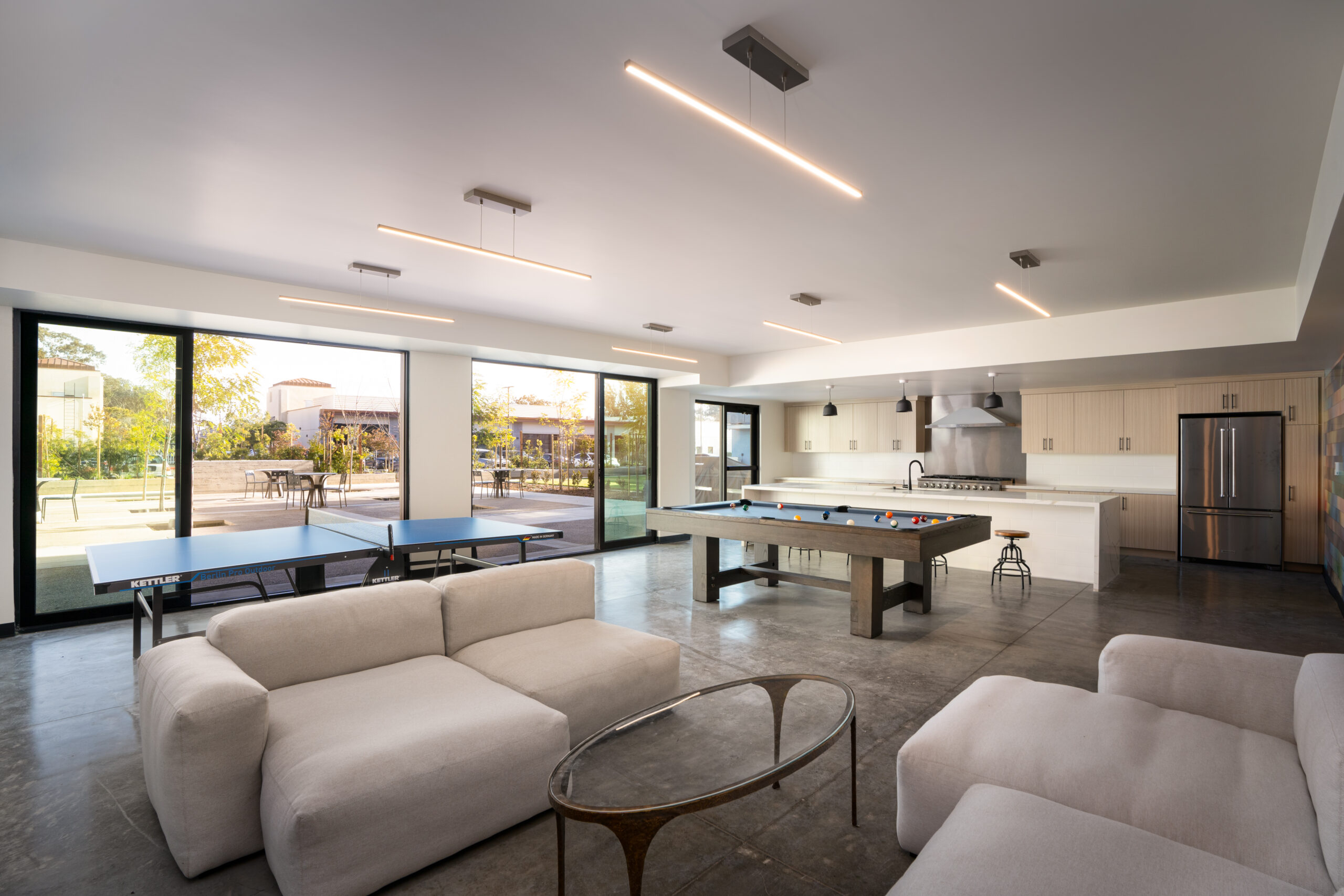 Interior architectural photograph of the common kitchen area from Elements Apartments in Santa Maria, California. A Pool table, ping pong table, stove, and triple wide and open floor to ceiling sliding glass doors are visible to capture an indoor-outdoor experience